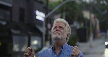 Yağmurda ayakta duran yaşlı adam, elleri açık ve duygusal ifade ile yukarı bakıyor keder, kafa karışıklığı, ya da ruhsal özlem şehir caddesinde