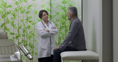 Muayene masasında oturan orta yaşlı erkek hasta klinikte kadın doktorla tartışıyor, sakin sağlık hizmetleri ortamında tıbbi danışmaya hazırlanıyor.