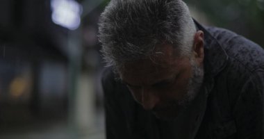 Orta yaşlı bir adam, başı yağmurda öne eğik, derin bir üzüntü ya da yorgunluk ifade ediyor.