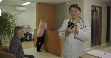 Doktorlar hastane lobisinde akıllı telefon kullanırken hastalar bekliyor ve hemşire arka planda asansörle modern tıbbi klinikte resepsiyona bakıyor.