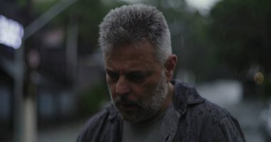 Orta yaşlı bir adam, ciddi bir şekilde aşağıya bakıyor, yağmurda sırılsıklam, hüzünlü bir yansıma ve duygusal ağırlık ifade ediyor.