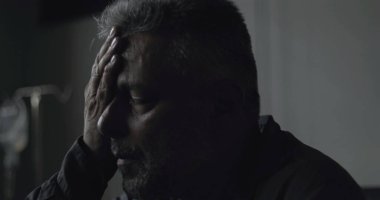 Klinikte gözleri kapalı oturan orta yaşlı bir adamın yakın plan çekimi, tıbbi belirsizlik anında sessizce ağrıyı ve yorgunluğu işliyor.