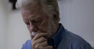 Son sınıf öğrencisi, düşünceli bir ifadeyle çenesini dinlendirirken, derin düşüncelere dalmış, sessiz bir odada aşağıya bakıyor.