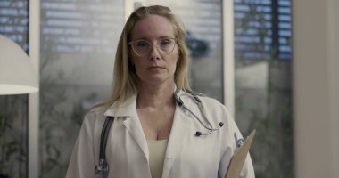 Ciddi bir kadın doktor panoyu tutuyor ve profesyonel tıp bürosundaki geniş pencereli ve kapalı alandaki bitkilere bakıyor.