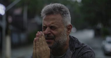 Orta yaşlı, gözleri kapalı ve elleri umutsuzca dua eden ya da duygusal yalvaran yağmurda ayakta duran bir adam.