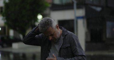 Orta yaşlı bir adam, yağmurun altında, elleri başının üstünde, gözleri açık, hayal kırıklığı ve zihinsel yorgunluk içinde.