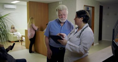 Kadın doktoru dinlerken gülümseyen kıdemli adam klinik lobisindeki dijital tablet hakkında bilgi veriyor, resepsiyon masasının yanındaki günlük sağlık danışmanlığı