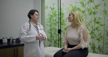 Doktor, yeşil bambu bitkisi geçmişi olan klinikteki sağlık danışmanlığı sırasında muayene masasında oturan orta yaşlı kadın hastayla konuşuyor.