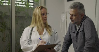 Doktor ve orta yaşlı erkek hasta, klinikte muayene sırasında arka planda pano ve tıbbi ekipmanlarla tartışıyorlar.