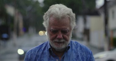 Yaşlı bir adam yağmurun altında üzgün gözlerle ve duygusal ifadeyle, dudakları sımsıkı ve kasvetli bir şekilde duruyor.