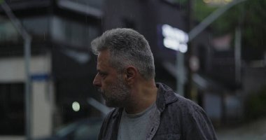 Orta yaşlı bir adam endişeli bir ifadeyle, gergin duruşu ve şehir sokaklarında duygusal ağırlığıyla yan yana bakıyor.