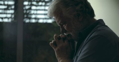 Derin yansıtıcı dualarda el ele tutuşan yaşlı bir adam, yumuşak ışık ve kasvetli atmosferiyle pencerenin yanında sessizce duruyor.