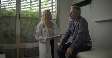 Klinikteki muayene masasında oturan orta yaşlı erkek hastayla tıbbi notları tartışırken gülümseyen kadın doktor.