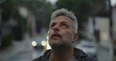 Orta yaşlı bir adam yağmurda dalgın dalgın bir ifadeyle yukarı bakıyor, boş şehir sahasında öylece duruyor.