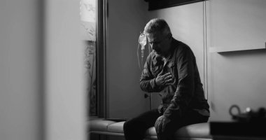 Klinikte başı dertteyken oturan orta yaşlı bir adam, dramatik siyah beyaz görüntü, tıbbi çevrede duygusal acı ya da zihinsel yük taşıyor.