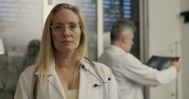 Steteskoplu kendine güvenen kadın doktor modern tıp bürosundaki kameraya bakarken erkek meslektaşı arka plandaki röntgeni inceliyor.