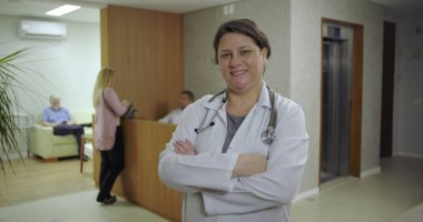 Klinik koridorunda kolları çapraz gülümseyen, steteskop ve laboratuvar önlüğü giyen, resepsiyondaki ve arka gruptaki hastalar tarafından görülebilen kendinden emin bir kadın doktor.