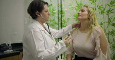 Klinikteki orta yaşlı kadın hastanın kalbini incelemek için steteskop kullanan kadın doktor rutin kontrolleri ve kişiselleştirilmiş tıbbi arabaları vurguluyor.