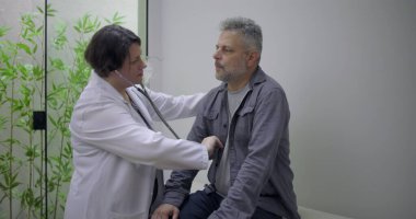 Steteskop kullanan kadın doktor klinik ortamdaki rutin sağlık kontrolleri sırasında orta yaşlı erkek hastaların kalp atışlarını inceliyor.