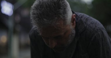 Orta yaşlı bir adam, başı yağmurda öne eğik, derin bir üzüntü ya da yorgunluk ifade ediyor.
