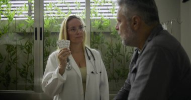 Tıbbi konsültasyon sırasında ilgili erkek hastaya reçeteli ilaçları açıklayan kadın doktor klinik ortamında su toplayan haplar tutuyor.
