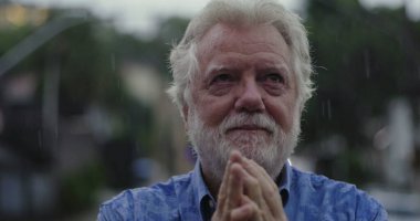 Yüzüne yapışmış elleri tutan yaşlı bir adam, yağmurda parlayan gözler, duygu, umut, ya da ruhsal bağlantı anında sessiz dua ile dolu.