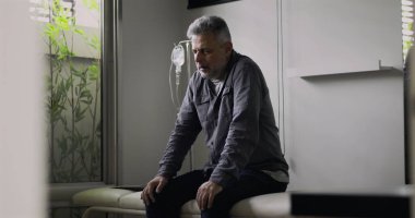 Orta yaşlı bir adam, boş bir muayenehanede gergin bir duruşla öne eğiliyor. Zor bir muayeneden sonra, yüzündeki gerginlik ve yorgunluk görülebiliyor.