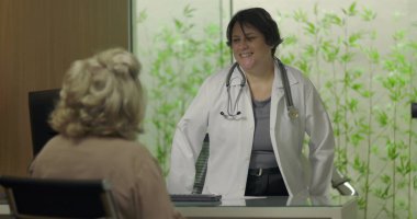 Steteskoplu gülümseyen doktor profesyonel klinikteki tıbbi danışmanlık sırasında yaşlı kadın hastayla samimi bir şekilde temas kuruyor.