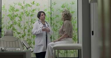 Klinikteki tıbbi danışmanlık sırasında muayene masasında oturan yaşlı kadın hastaya el hareketleri ve profesyonel odaklanma ile teşhisi açıklayan doktor.