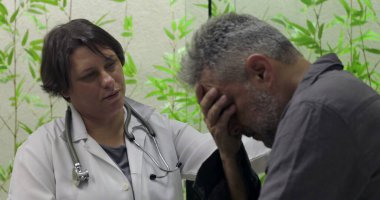 Sıkıntılı orta yaşlı erkek hasta yüzünü kapatırken, kadın doktor klinikte destek sunuyor, zor tıbbi haberlere duygusal ifadeyle tepki veriyor.