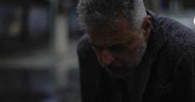 Orta yaşlı bir adam, başı yağmurda öne eğik, derin bir üzüntü ya da yorgunluk ifade ediyor.