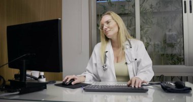 Tıp bürosunda masaüstü bilgisayarı üzerinde çalışan, hasta veri girişine odaklanan ya da klinik çalışma oturumunda rapor yazan kadın doktor