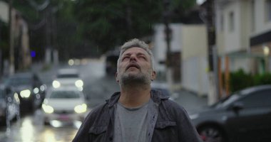 Orta yaşlı bir adam yağmurda dikiliyor, başı arkaya eğik ve gözleri kapalı, rahatlama, üzüntü veya duygusal katarsi ifade ediyor.