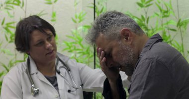 Sıkıntılı orta yaşlı erkek hasta yüzünü kapatırken, kadın doktor klinikte destek sunuyor, zor tıbbi haberlere duygusal ifadeyle tepki veriyor.