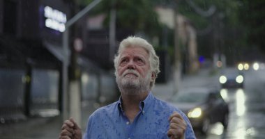 Yağmurda ayakta duran yaşlı adam, elleri açık ve duygusal ifade ile yukarı bakıyor keder, kafa karışıklığı, ya da ruhsal özlem şehir caddesinde