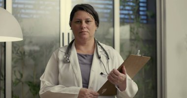 Doktor ofisinde elinde bir panoyla duruyor ve modern klinik ortamda tarafsız bir ifadeyle kameraya bakıyor.