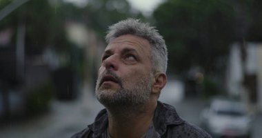 Orta yaşlı bir adam gökyüzüne doğru bakıyor hüznün ya da özlemin açık ifadesi, yağmurda hareketsiz duruyor.