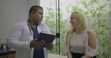 Klinik ziyareti sırasında orta yaşlı kadın hastaya tıbbi bilgileri açıklamak için tablet kullanan erkek doktor, sağlık hizmetlerinde güveni ve netliği artırıyor