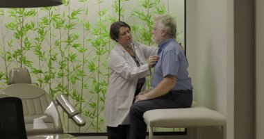 Yaşlı erkek hasta üzerinde steteskopla göğüs muayenesi yapan kadın doktor klinikteki tıbbi muayene masasında oturuyor.