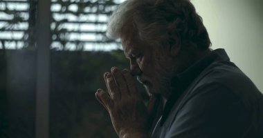 Derin yansıtıcı dualarda el ele tutuşan yaşlı bir adam, yumuşak ışık ve kasvetli atmosferiyle pencerenin yanında sessizce duruyor.