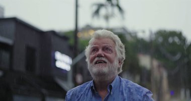 Kolları geniş ve yüzü göğe yükselmiş yaşlı bir adam, yağmuru ruhsal rahatlama, minnettarlık ya da duygusal teslimiyet ifadesiyle kucaklıyor.