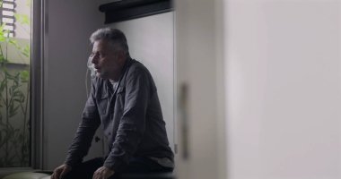 Orta yaşlı bir adam, boş bir muayenehanede gergin bir duruşla öne doğru eğiliyor. Zor bir muayeneden sonra, dışavurumunda görülen gerginlik ve yorgunluğu yansıtıyor.