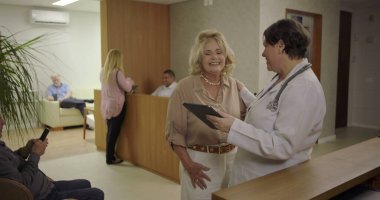 Klinikte resepsiyon masasının yanında kadın doktorla dijital tablet üzerinde tıbbi bilgileri inceleyen yaşlı bir kadın, ciddi sağlık sorunları