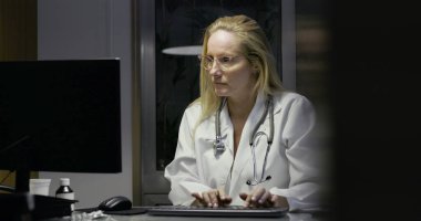 Masada geç saatlere kadar çalışırken not alan kadın doktor, tıbbi malzemeler ve belgelerle çevrili, odaklanmış ifadeli bilgisayar monitöründen uzağa bakıyor.