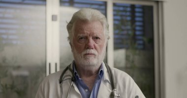 Beyaz sakallı ve dosyalı deneyimli bir erkek doktor klinikte dikilip odaklanmış ekspresiyoyla doğrudan kameraya bakıyor.