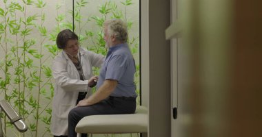 Kadın doktor göğüs muayenesi yaparken gülümsüyor. Yaşlı erkek hasta üzerinde steteskopla klinikte oturmuş yeşil yapraklı waffle ile süslenmiş.