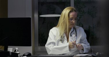 Masada geç saatlere kadar çalışırken not alan kadın doktor, tıbbi malzemeler ve belgelerle çevrili, odaklanmış ifadeli bilgisayar monitöründen uzağa bakıyor.