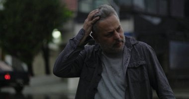 Orta yaşlı bir adam, yağmurun altında, elleri başının üstünde, gözleri kapalı, hayal kırıklığı ya da zihinsel yorgunluk içinde.