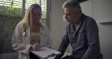 Hasta muayene masasında oturan orta yaşlı bir erkeğe danışmanlık yaparken elinde dosya tutan kadın doktor.