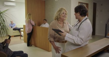 Kliniğin resepsiyonunda kadın doktorun tuttuğu dijital tablete bakan yaşlı kadın, güven ve dikkat ile günlük sağlık hizmetleri sohbeti